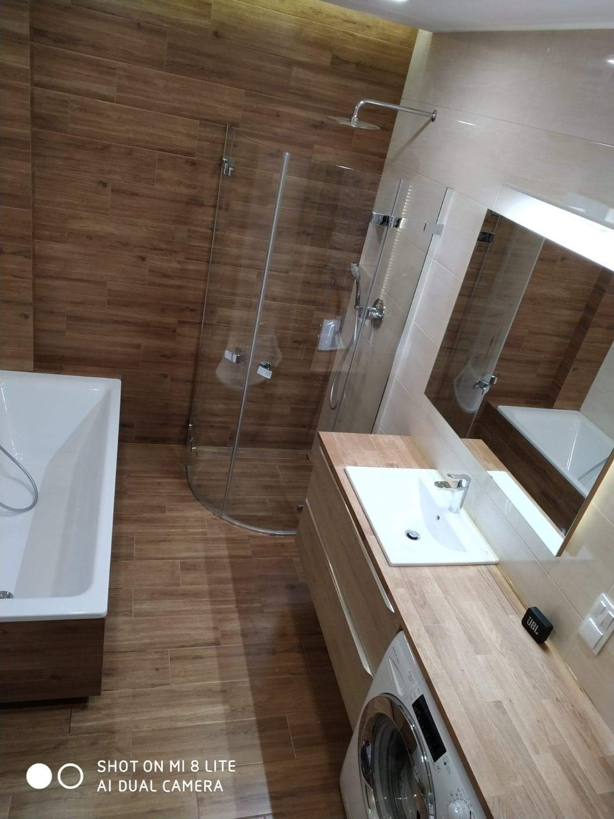 Nowoczesna łazienka z drewnopodobnymi płytkami, kabiną prysznicową typu walk-in, białą wanną, umywalką nablatową na drewnianym blacie i pralką pod blatem.