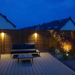 Oświetlony ogród wieczorową porą z drewnianym tarasem, meblami ogrodowymi i podświetlonym drzewem na tle drewnianego ogrodzenia z lampami.