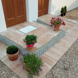 Wykończone wejście do domu z beżowej kostki brukowej, obrzeża z szarej kostki, donice z kwiatami i krzewami ozdobnymi.