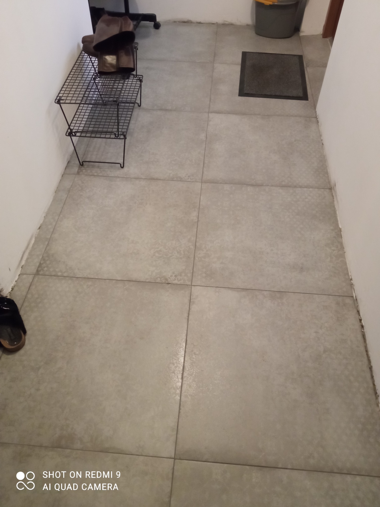 Szare płytki podłogowe ułożone w kwadraty, widoczna metalowa półka na buty z parą butów, wycieraczka i wiadro w rogu pomieszczenia.