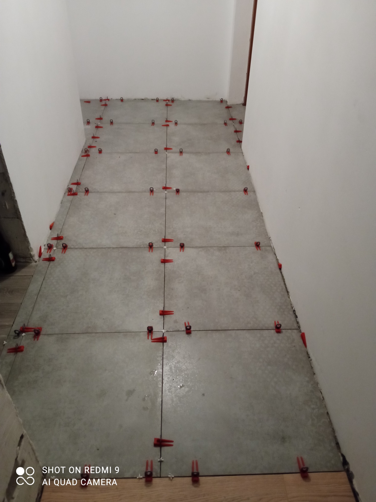 Szare płytki podłogowe w trakcie układania, z czerwonymi klipsami poziomującymi spoiny, widoczne fragmenty ściany i podłogi z drewna.