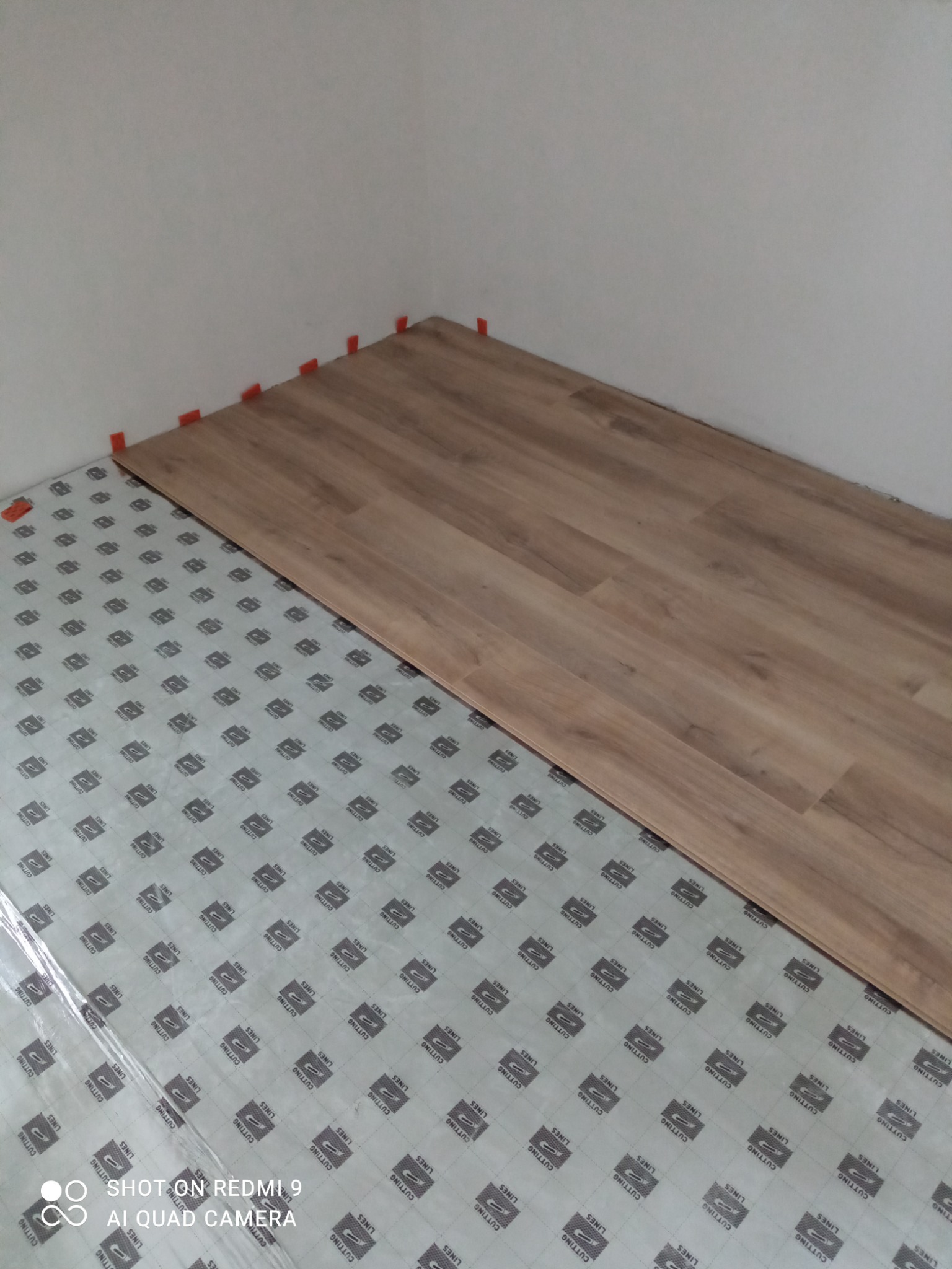 Fragment podłogi w trakcie układania parkietu, widoczne panele i podkład z nadrukiem, pomarańczowe kliny dystansowe przy ścianie.