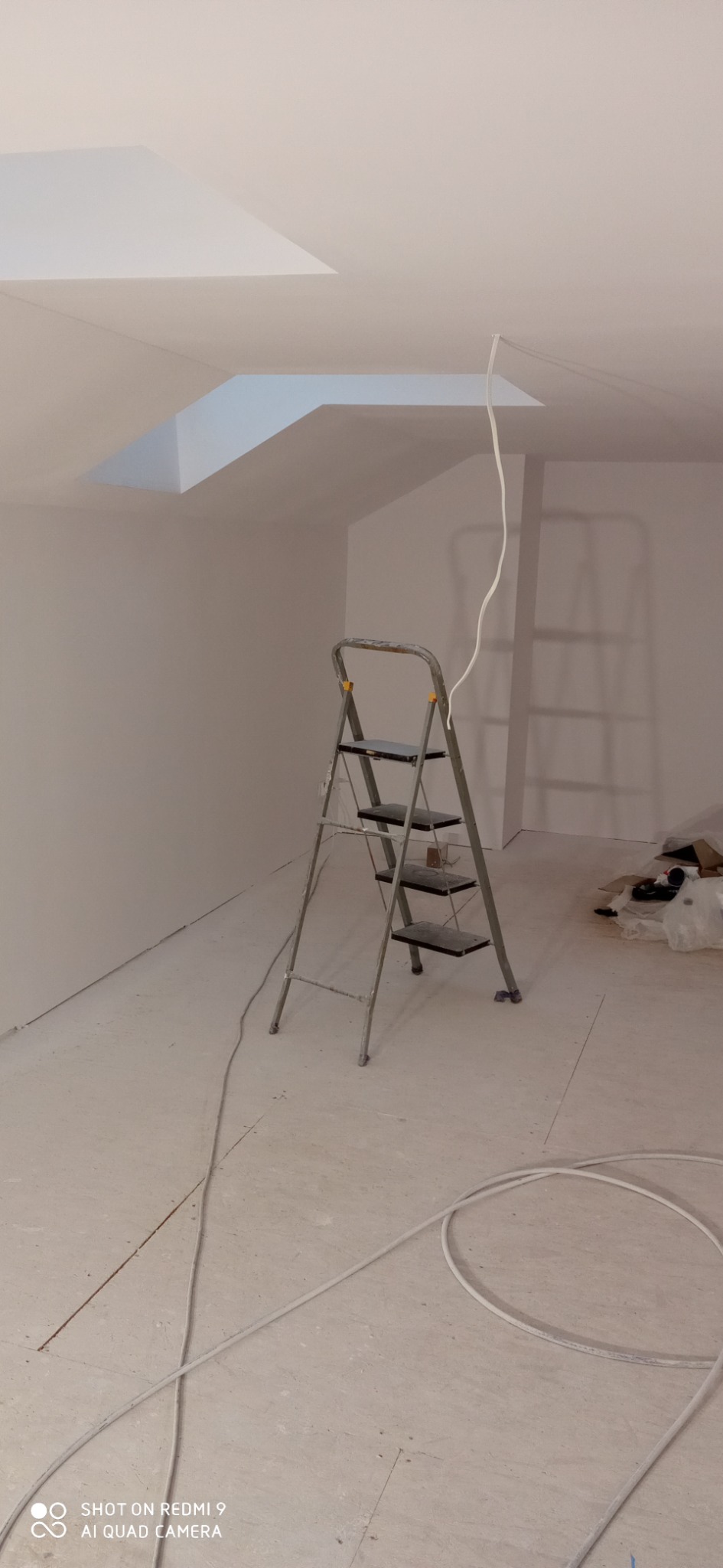 Pokój w Koszalinie w trakcie remontu, widoczna drabina aluminiowa, białe ściany po gładziowaniu, kabel zwisający z sufitu i resztki materiałów budowlanych.