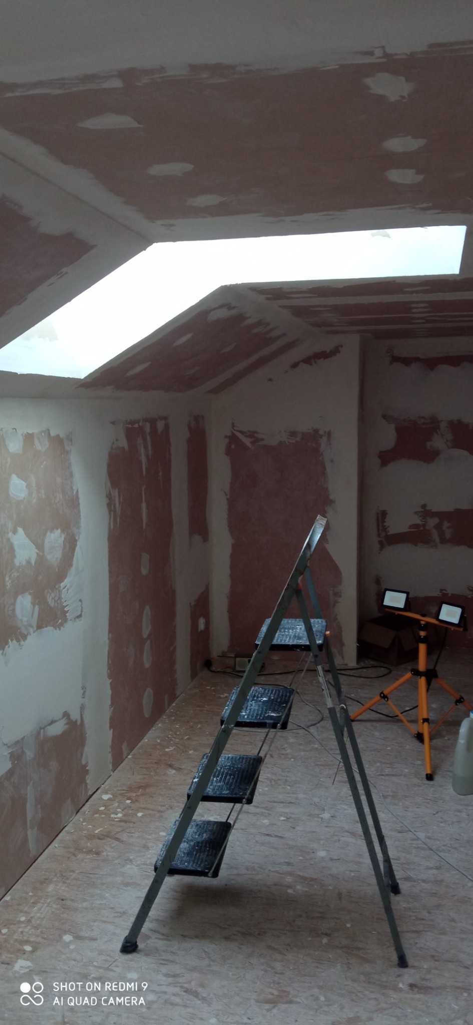Wnętrze w trakcie wykańczania, ściany i sufit pokryte warstwą szpachli, widoczne ślady po łatowaniu, drabina aluminiowa i lampa budowlana na trójnogu w pomieszczeniu z oknem dachowym.