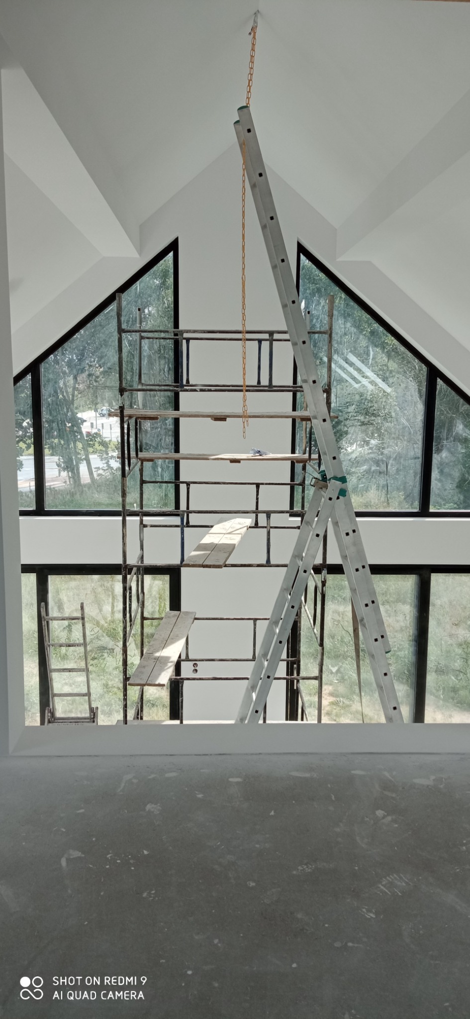 Wnętrze w trakcie remontu z rusztowaniem i drabiną na tle dużych okien w kształcie trójkątów, widok z góry na szarą podłogę.