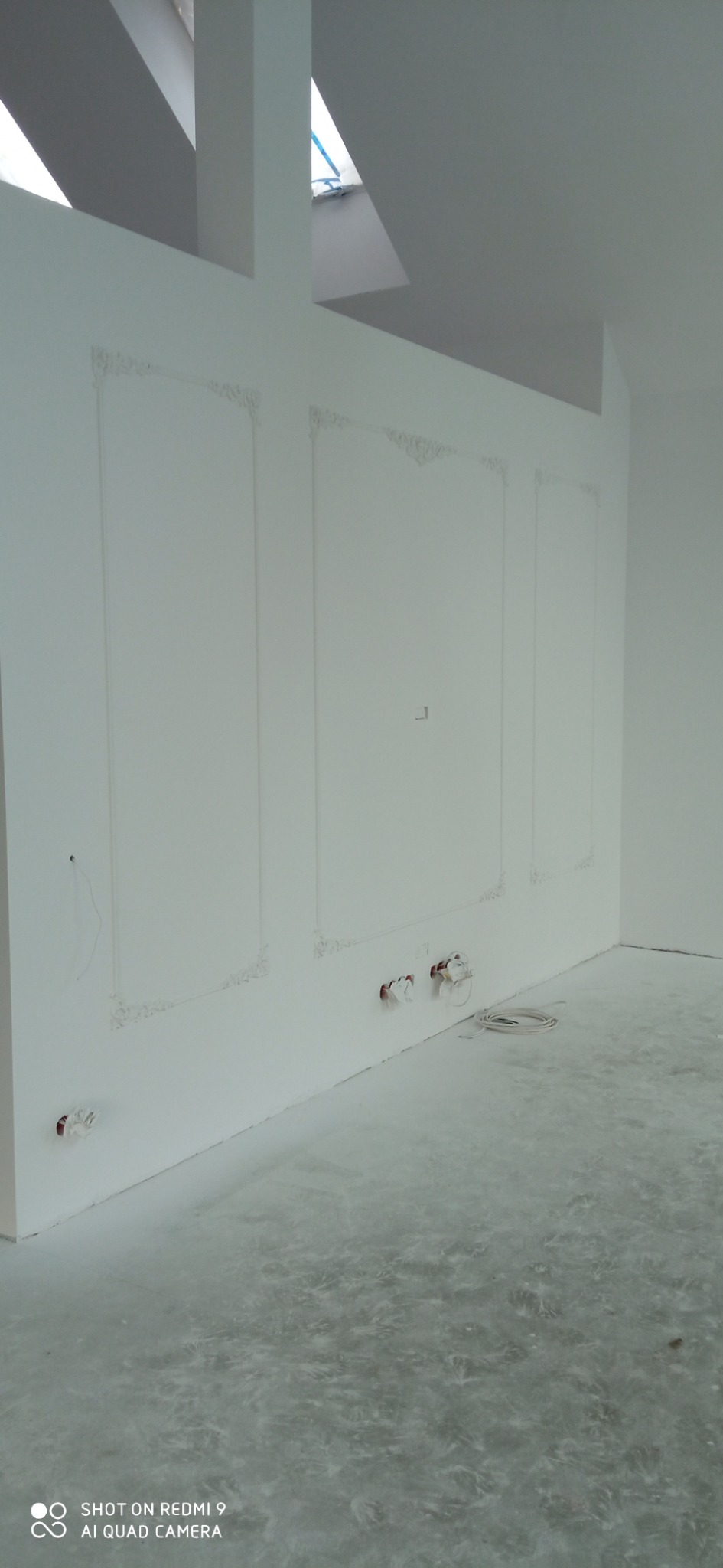 Świeżo położona, jasnoszara gładź na podłodze w pustym, białym pomieszczeniu z dekoracyjnymi listwami ściennymi i wystającymi przewodami elektrycznymi.