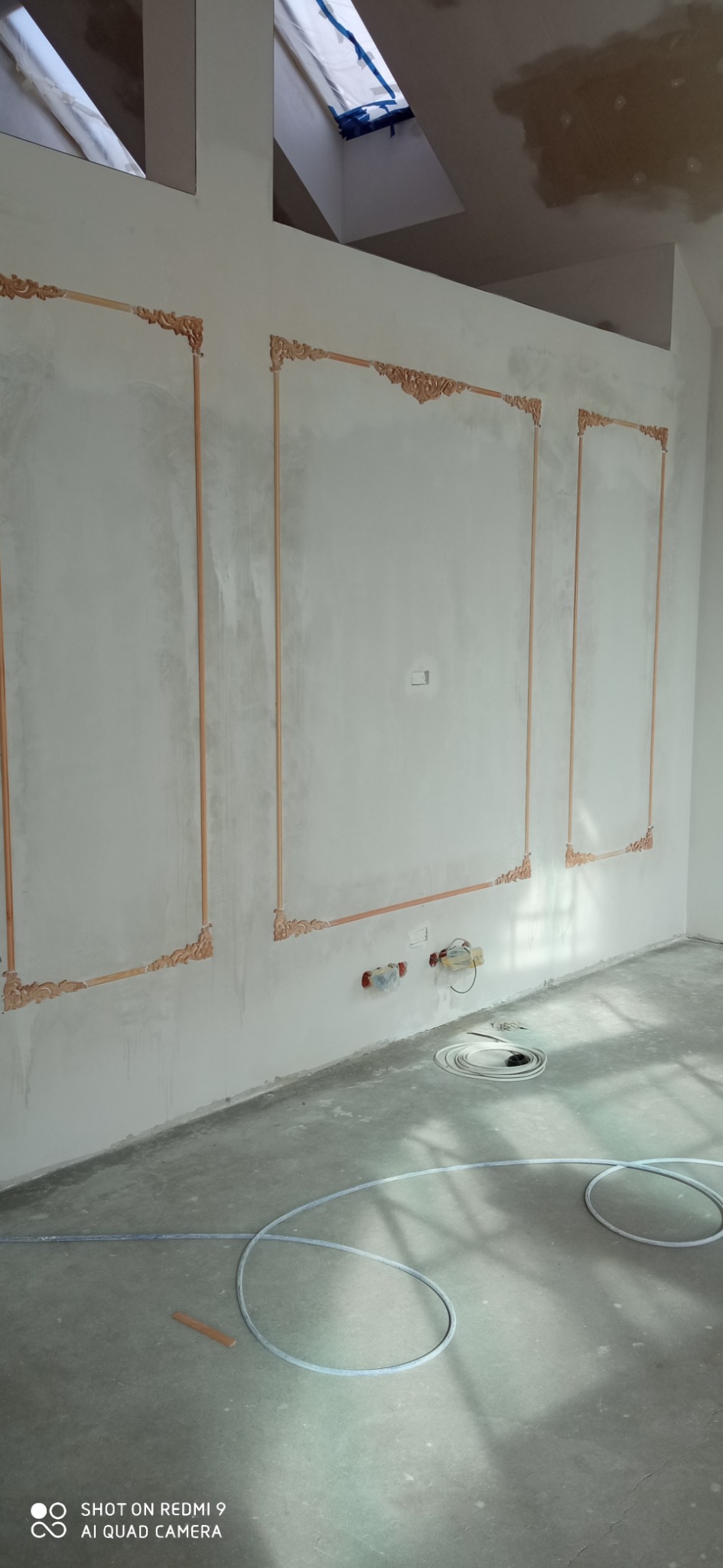 Ściana w trakcie wykańczania z dekoracyjnymi, drewnianymi listwami tworzącymi ramy, widoczne instalacje hydrauliczne i elektryczne, poddasze z oknem dachowym, surowy beton na podłodze i kable...