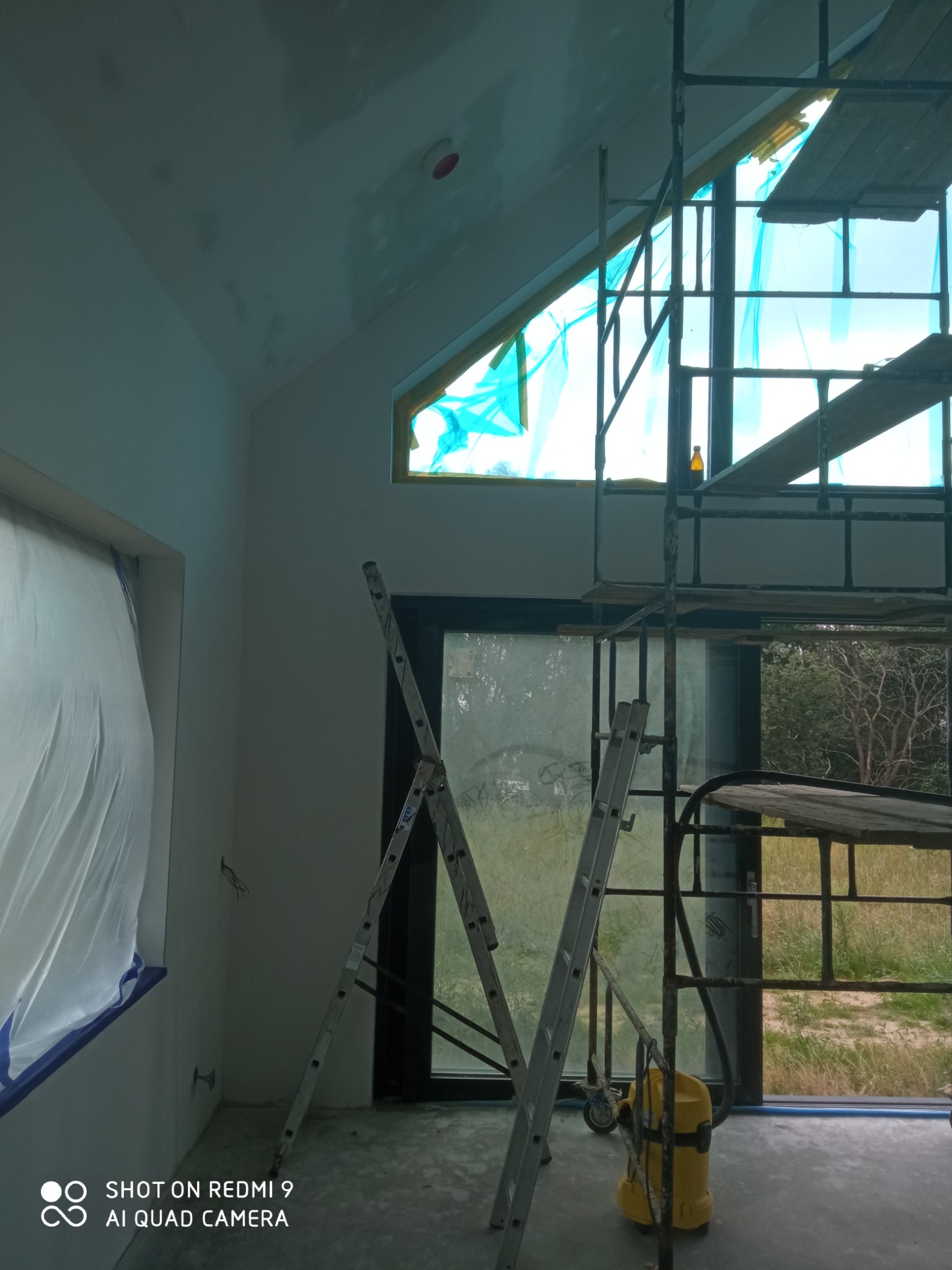 Wnętrze w trakcie remontu: ściany przygotowane do malowania, okna zabezpieczone folią, widoczne rusztowanie, drabina oraz odkurzacz przemysłowy.