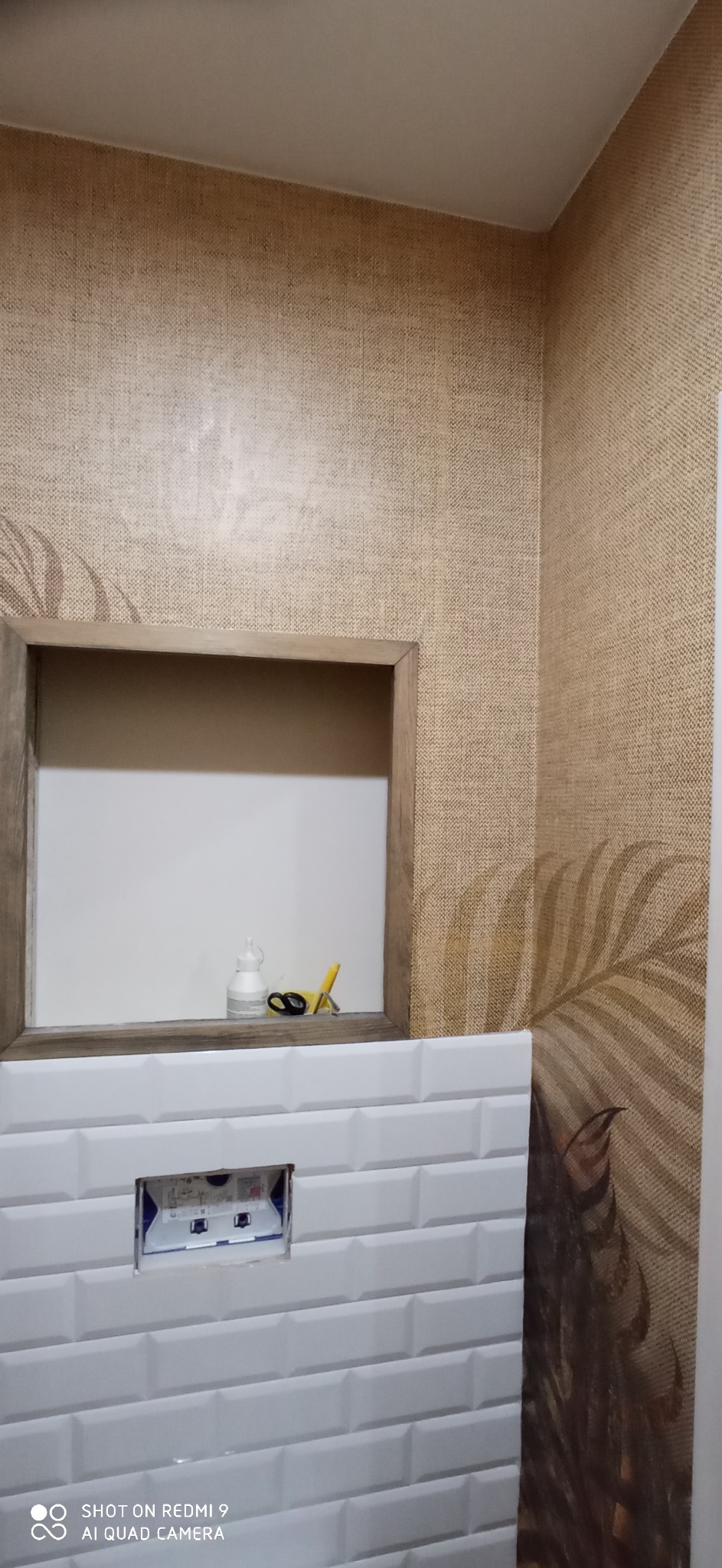 Ściana wykończona beżową tapetą z motywem liści w Koszalinie, z widoczną wnęką obudowaną drewnem, pod którą ułożono białe, prostokątne płytki, a w nich zamontowano stelaż podtynkowy do WC.