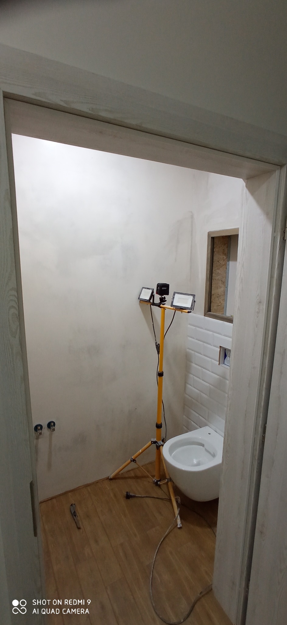 Wnętrze łazienki w trakcie remontu, ściana częściowo pokryta gładzią, widoczne przyłącza wodne, biała toaleta, oświetlenie budowlane na statywie, podłoga imitująca drewno.
