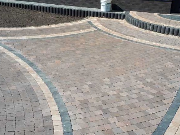 Ułożona kostka brukowa w odcieniach brązu i beżu, z dekoracyjnymi obrzeżami w kolorze szarym i beżowym, tworząca łukowaty wzór na podjeździe. W tle widoczna rabata z ziemią i mur z ciemnych cegieł...