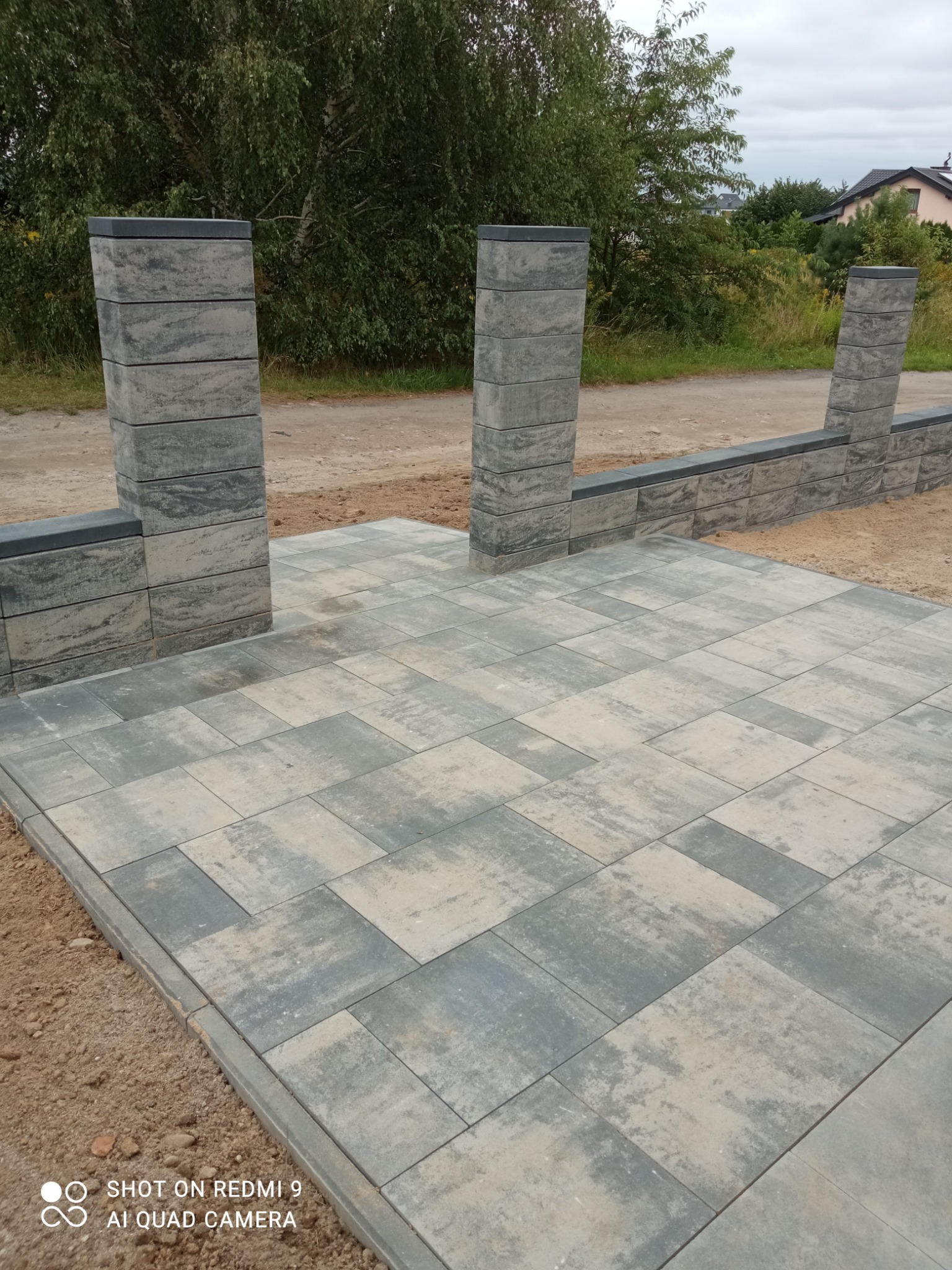 Ułożona kostka brukowa w Koszalinie, tworząca wzór szachownicy w odcieniach szarości i beżu, otoczona piaskiem i z elementami ogrodzenia z betonowych bloków. Widoczne słupki ogrodzeniowe z szarej...