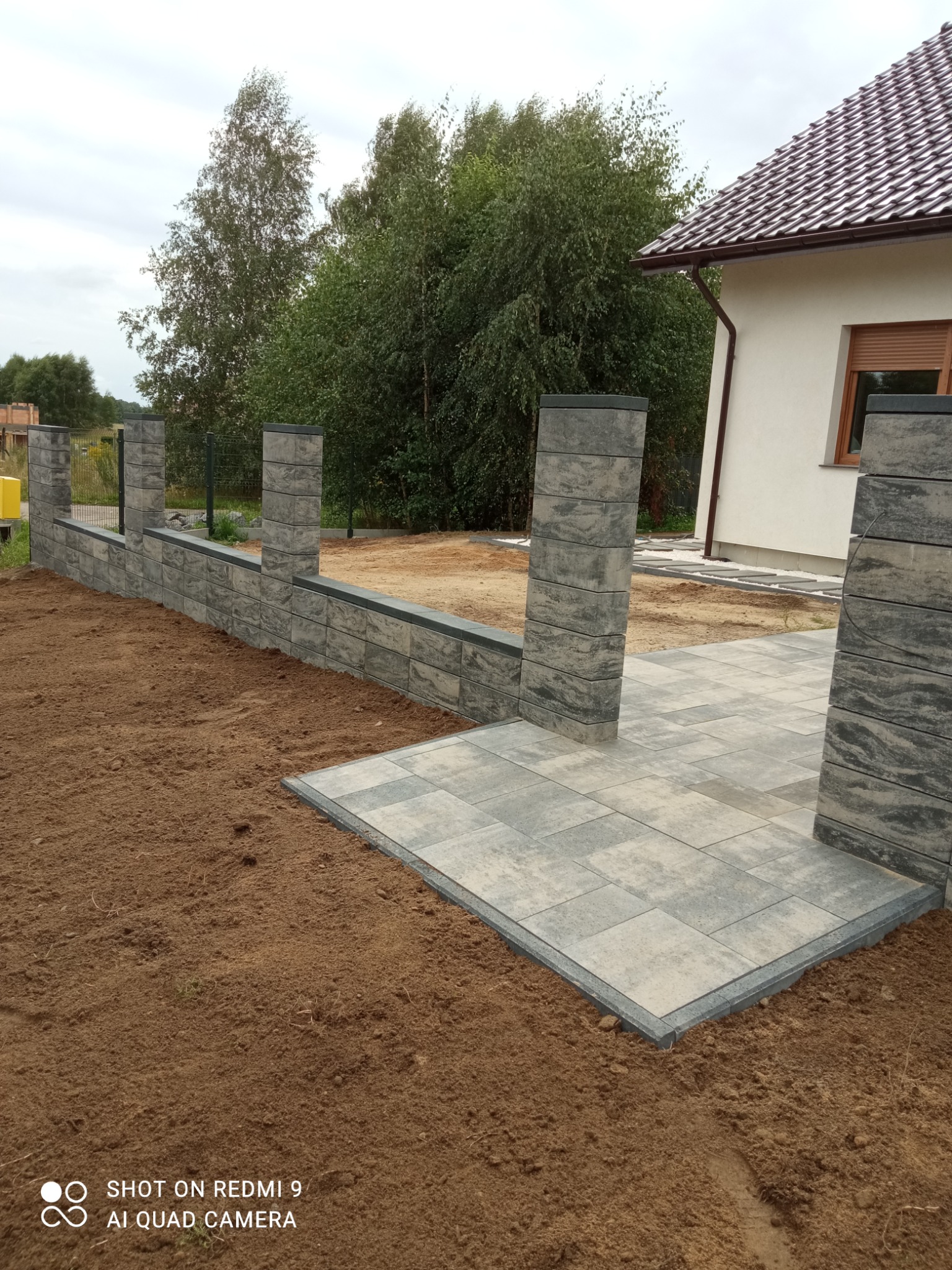 Nowo ułożona szara kostka brukowa tworząca taras i ścieżkę, otoczona przygotowanym pod trawnik podłożem, z widocznym fragmentem ogrodzenia z betonowych bloczków i domu z brązowym dachem w tle.