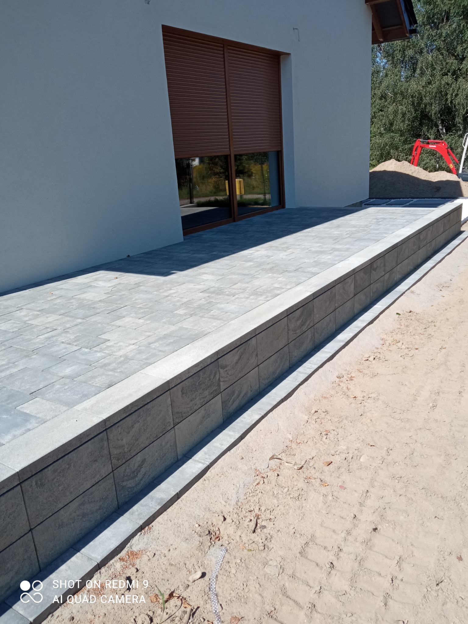 Nowoczesny podjazd z szarej kostki brukowej, wykończony obrzeżem z ciemniejszych, kwadratowych elementów. W tle widoczny fragment domu z brązowymi roletami i mini koparka na pryzmie piasku.