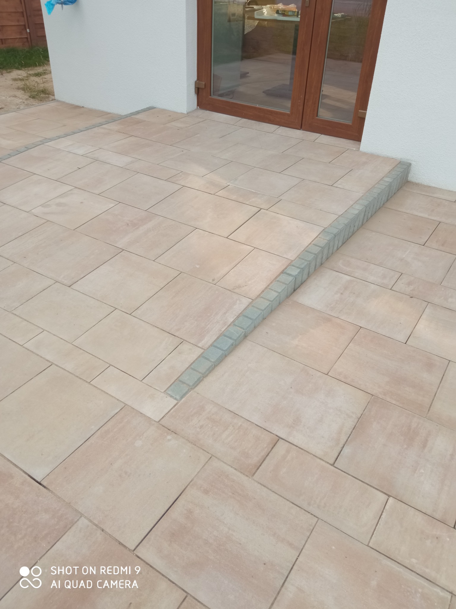 Ułożona beżowa kostka brukowa na tarasie przed domem, z szarym obramowaniem schodka przy drzwiach balkonowych.