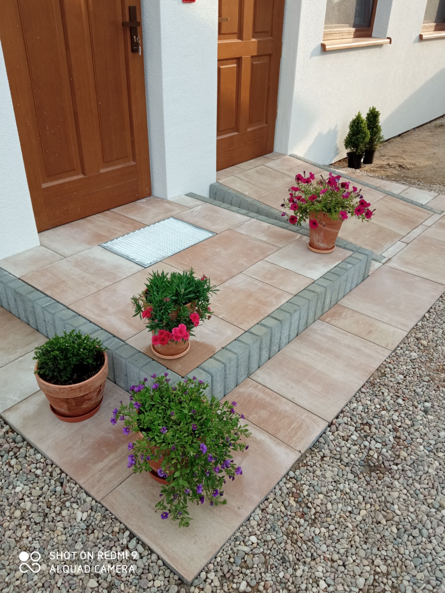 Wykończone wejście do domu z beżowej kostki brukowej, obrzeża z szarej kostki, donice z kwiatami i krzewami ozdobnymi.