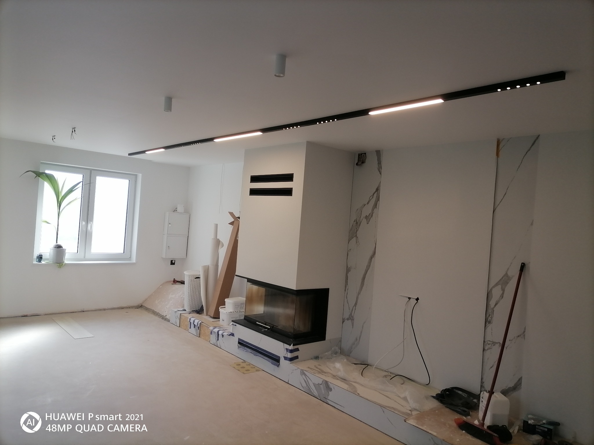 Wnętrze w trakcie prac wykończeniowych: zabudowany kominek z marmurowym wykończeniem, sufit z nowoczesnym oświetleniem liniowym, przygotowane podłoże pod posadzkę, widoczne narzędzia i materiały...