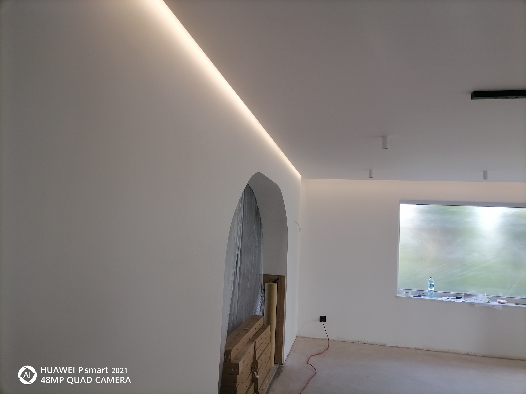 Wnętrze po szpachlowaniu i malowaniu na biało z oświetleniem LED w suficie, widoczny łuk drzwiowy, podłoga przygotowana do dalszych prac wykończeniowych, okno z butelką wody.