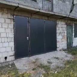 Trzyczęściowa, czarna brama przemysłowa zamontowana w murowanym budynku z białej cegły, widok z zewnątrz, zarośnięte podłoże.