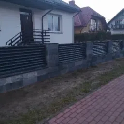 Nowoczesne ogrodzenie z poziomych, czarnych paneli i szarych, kamiennych słupków oraz podmurówki, oddzielające posesję od chodnika z czerwonej kostki.