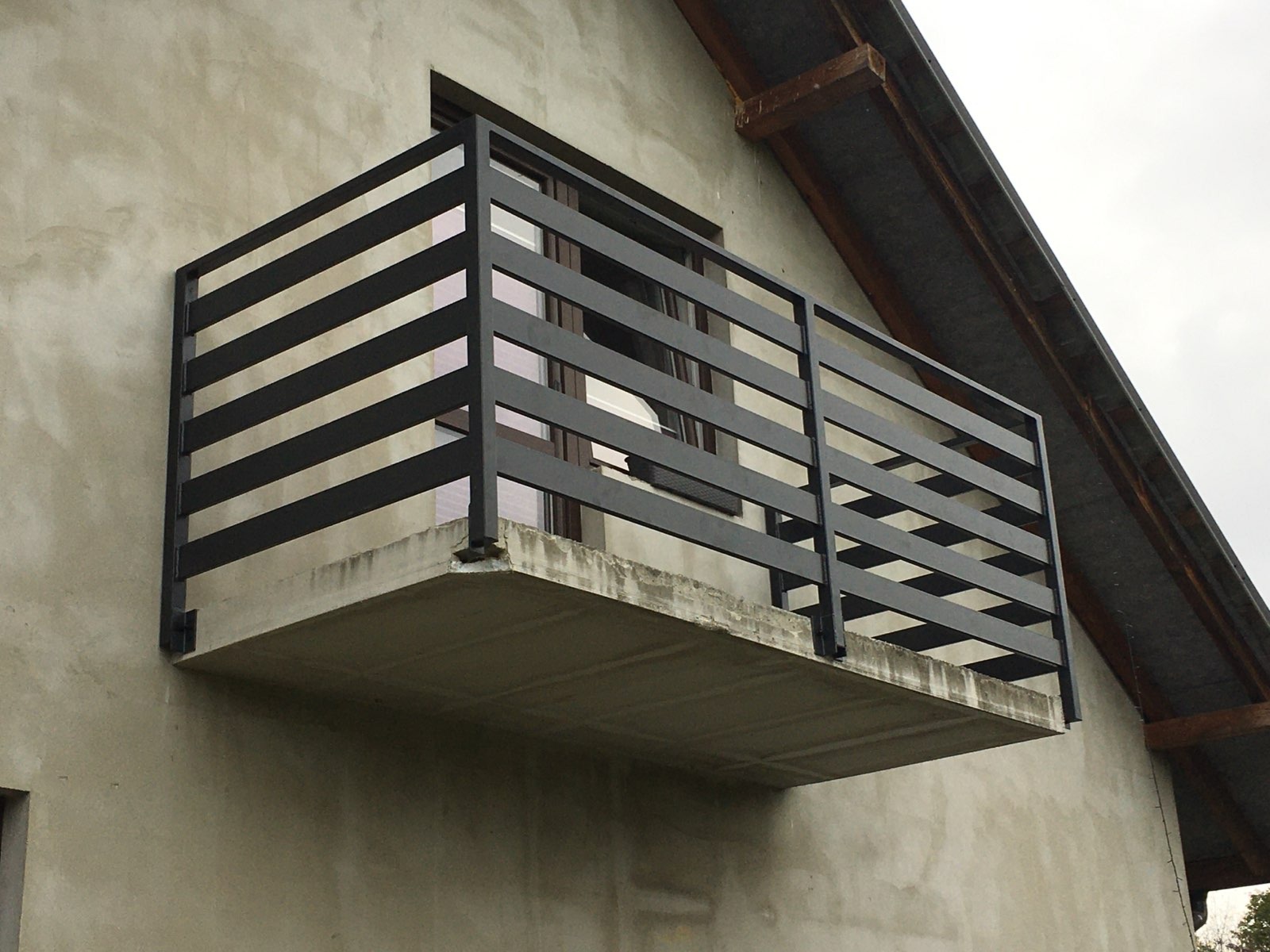 Nowoczesna, pozioma balustrada balkonowa z czarnych, metalowych profili na betonowej płycie balkonu, widok z dołu na tle szarej elewacji budynku.