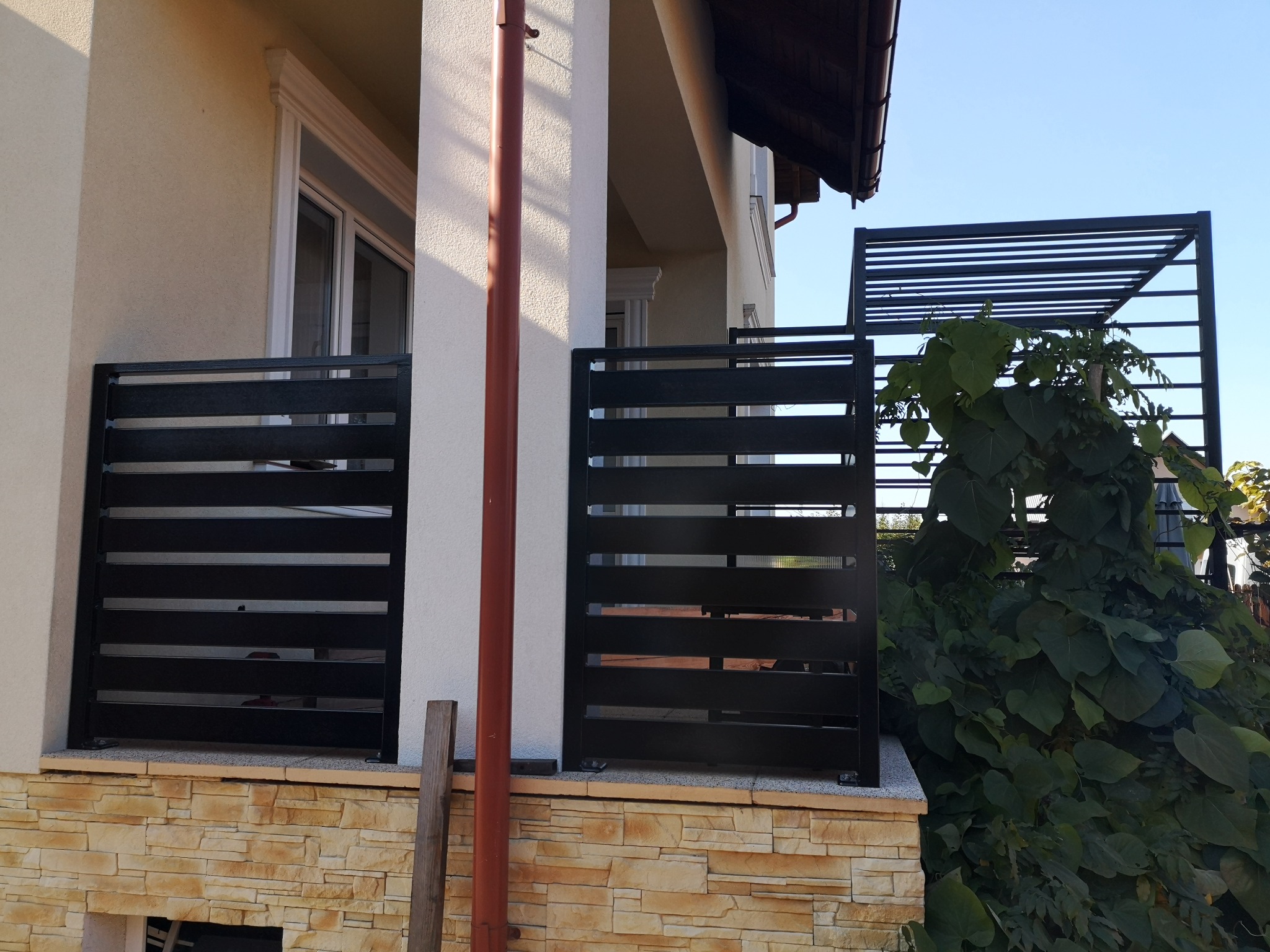 Nowoczesne ogrodzenie metalowe z poziomymi panelami w kolorze czarnym, zamontowane na murku z kamienia elewacyjnego, z widoczną konstrukcją pergoli w tle i zielenią pnącą się po niej.