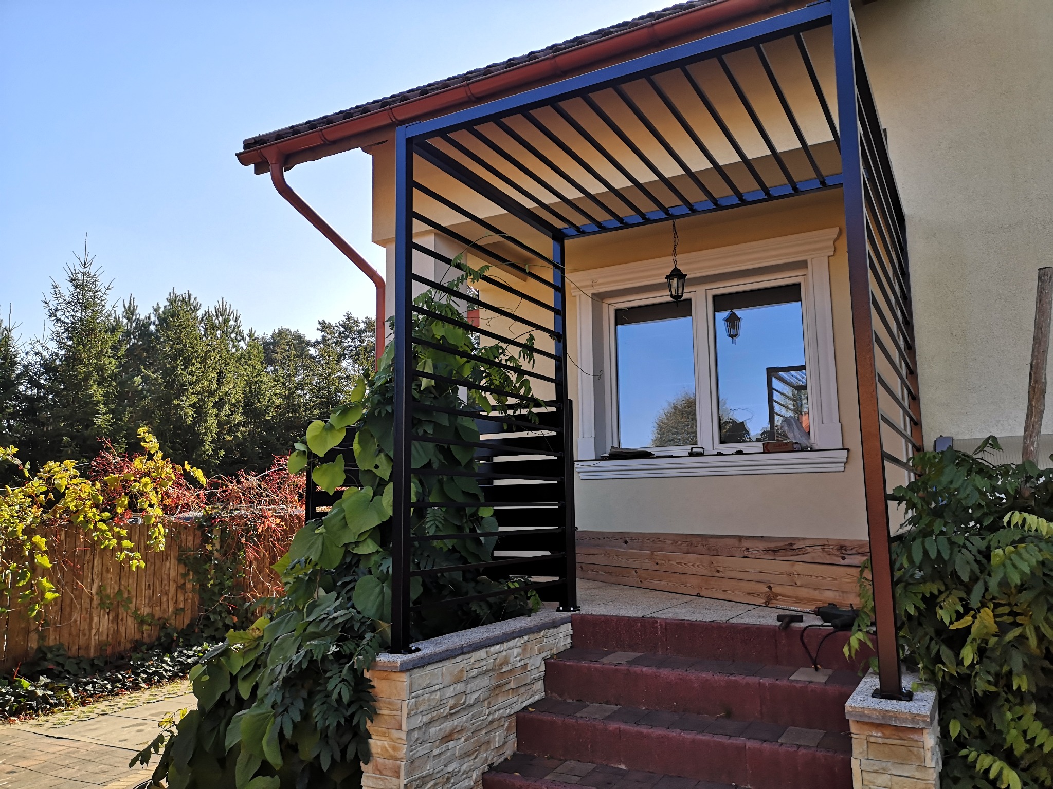 Czarna, nowoczesna konstrukcja z metalowych profili tworząca zadaszenie nad wejściem do domu, częściowo porośnięta zieloną roślinnością pnącą, widoczne schody wejściowe.