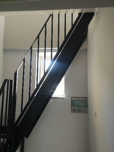 Czarne, metalowe schody z prostymi balustradami w białym wnętrzu, widok z dołu, okno w tle z odbijającym się światłem.