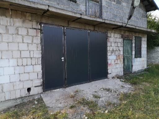 Trzyczęściowa, czarna brama przemysłowa zamontowana w murowanym budynku z białej cegły, widok z zewnątrz, zarośnięte podłoże.