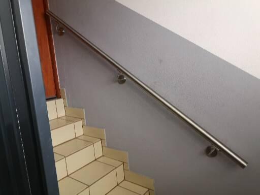 Wewnętrzne schody wyłożone jasnymi płytkami, z zamontowaną prostą, metalową poręczą przy ścianie o szarym odcieniu.