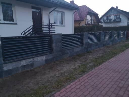 Nowoczesne ogrodzenie z poziomych, czarnych paneli i szarych, kamiennych słupków oraz podmurówki, oddzielające posesję od chodnika z czerwonej kostki.