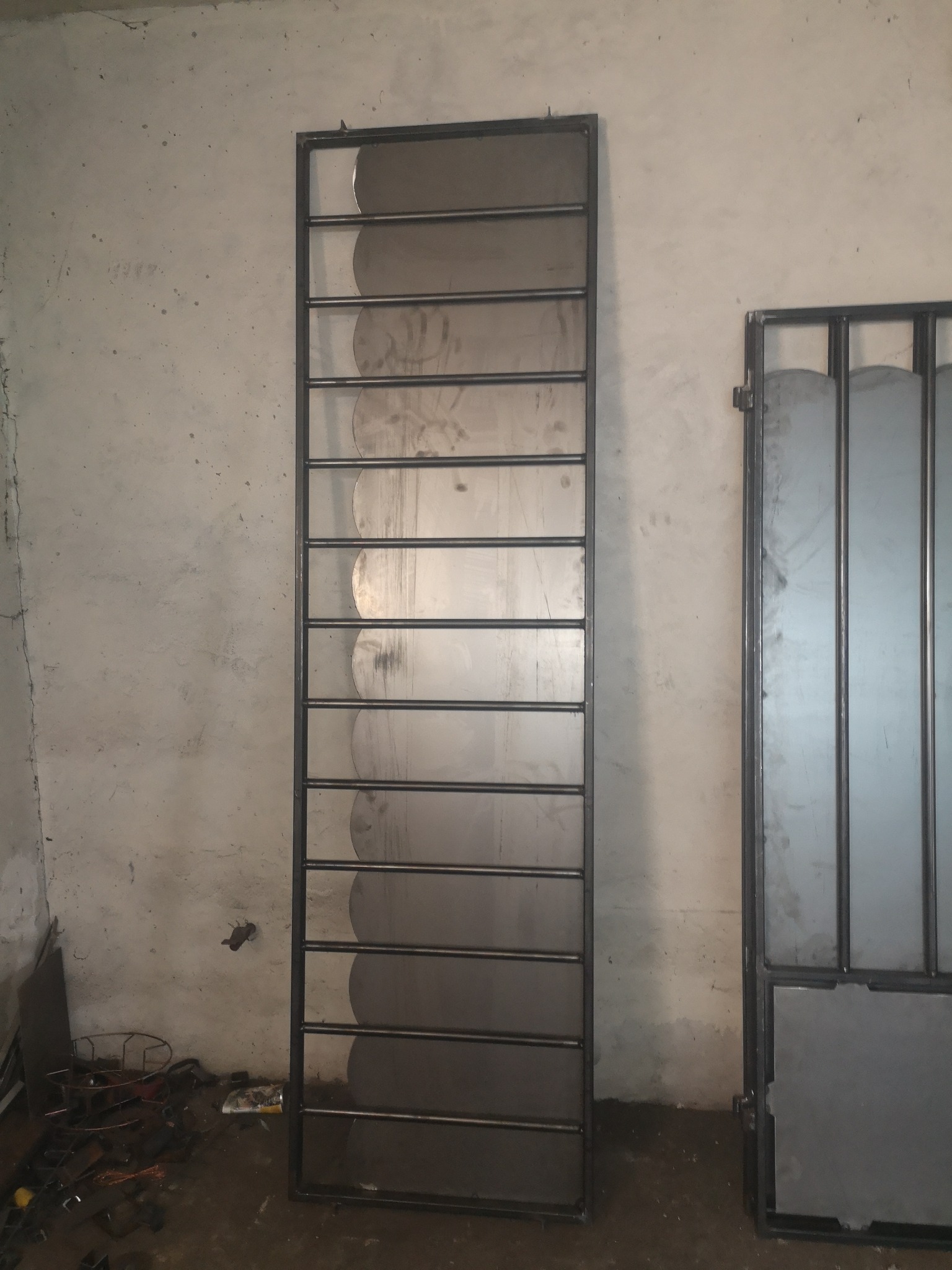 Surowa, metalowa konstrukcja kraty z zaokrąglonymi elementami wewnętrznymi, oparta o ścianę w warsztacie, obok częściowo widoczna rama bramy.