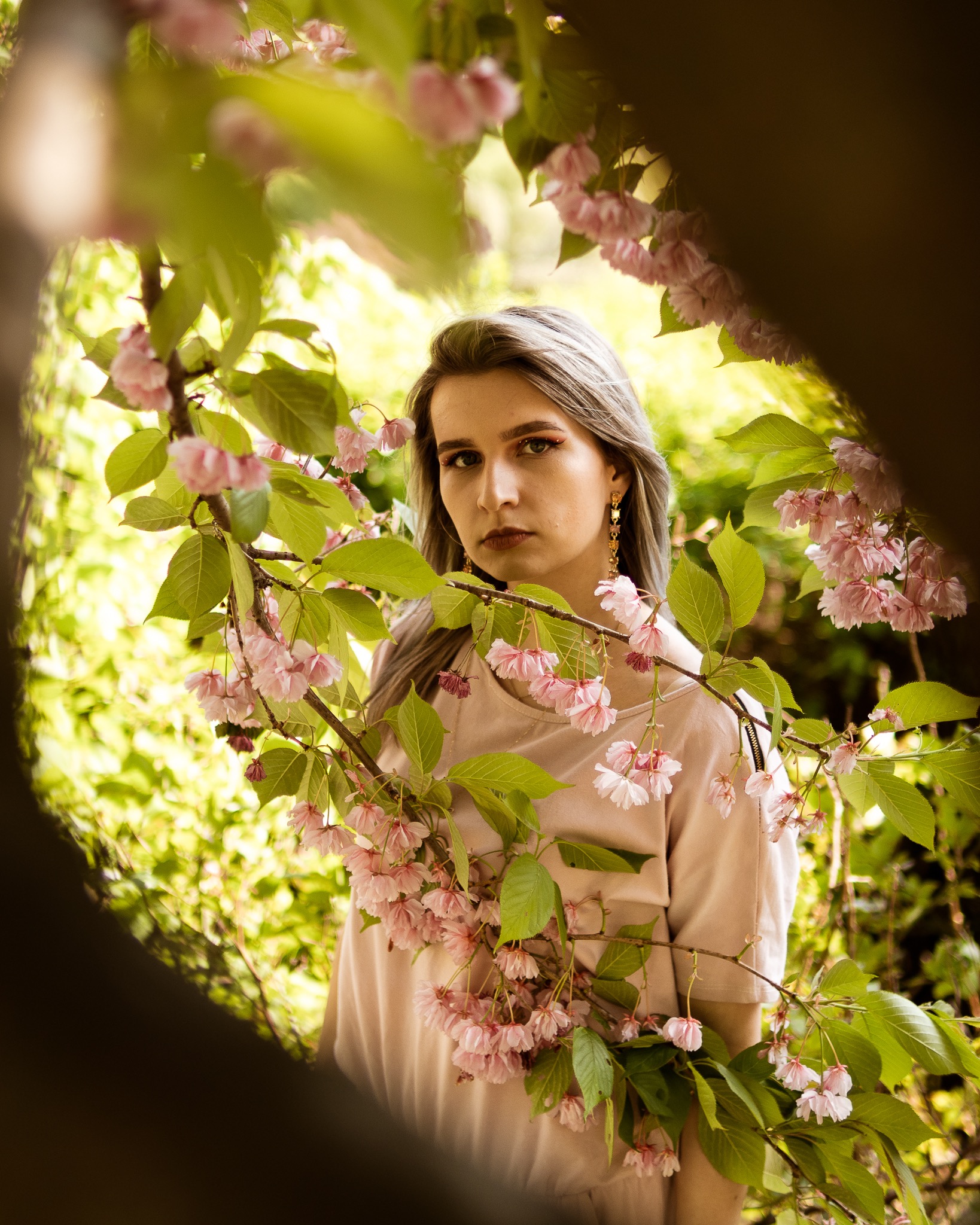 Portret kobiety z popielatymi włosami, pozującej w plenerze wśród kwitnącej wiśni, z gałązką kwiatów na pierwszym planie, ujęcie zza drzewa.