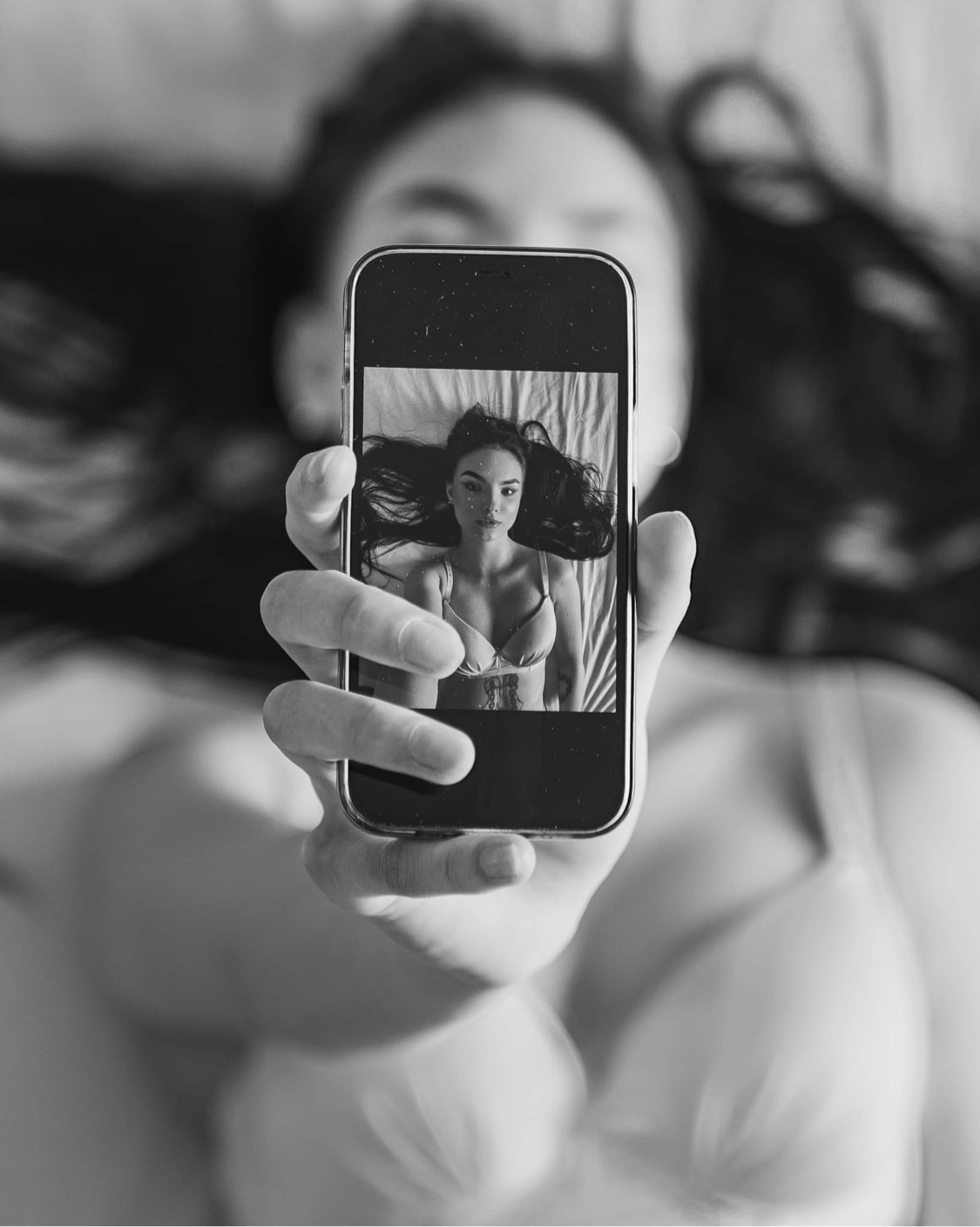 Czarno-białe zdjęcie kobiety trzymającej telefon, na ekranie którego widnieje jej portret w bieliźnie leżącej na łóżku, z widocznym tatuażem na żebrach.