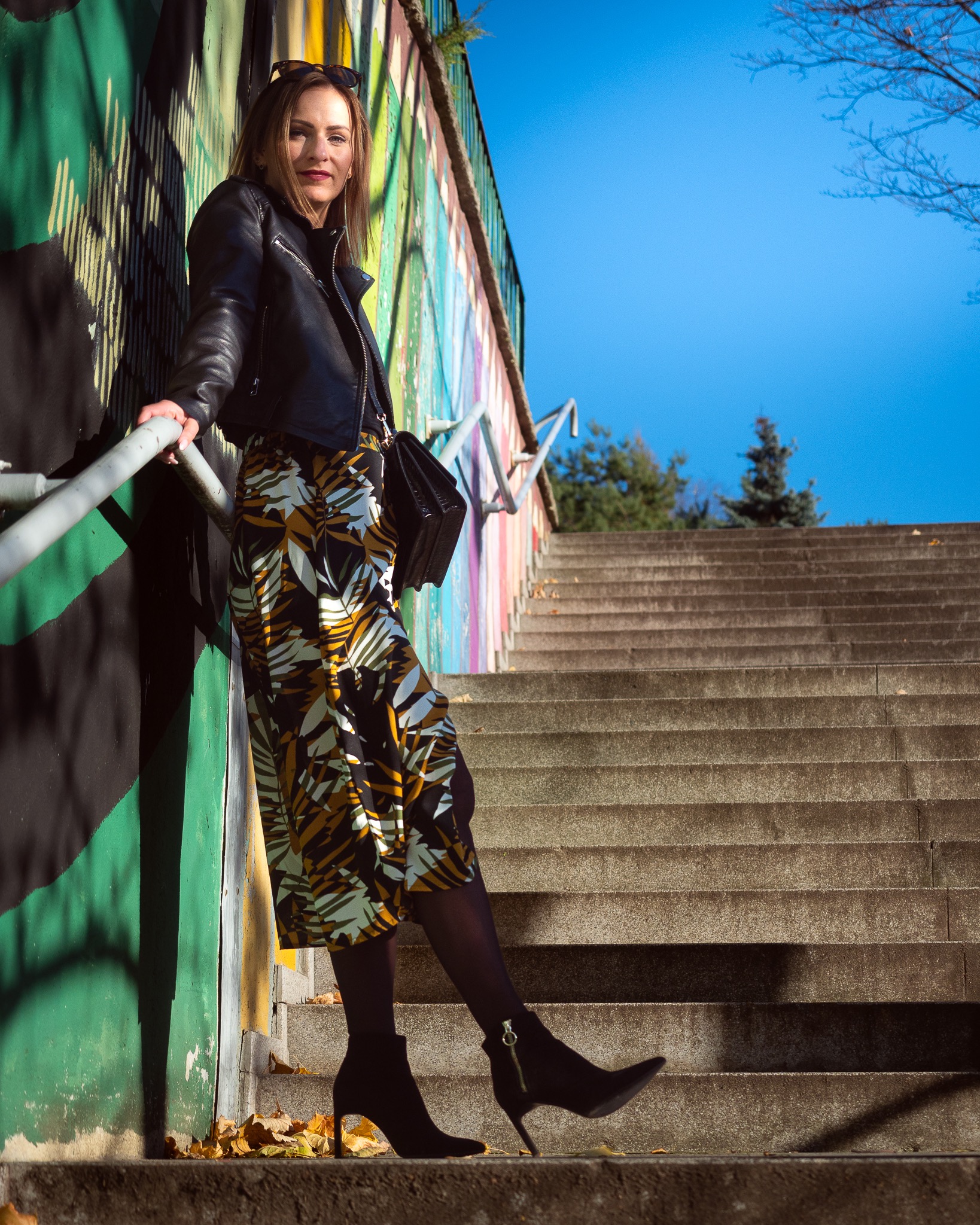 Kobieta w skórzanej kurtce i wzorzystych spodniach pozuje na tle graffiti i schodów, promienie słoneczne padają na twarz.