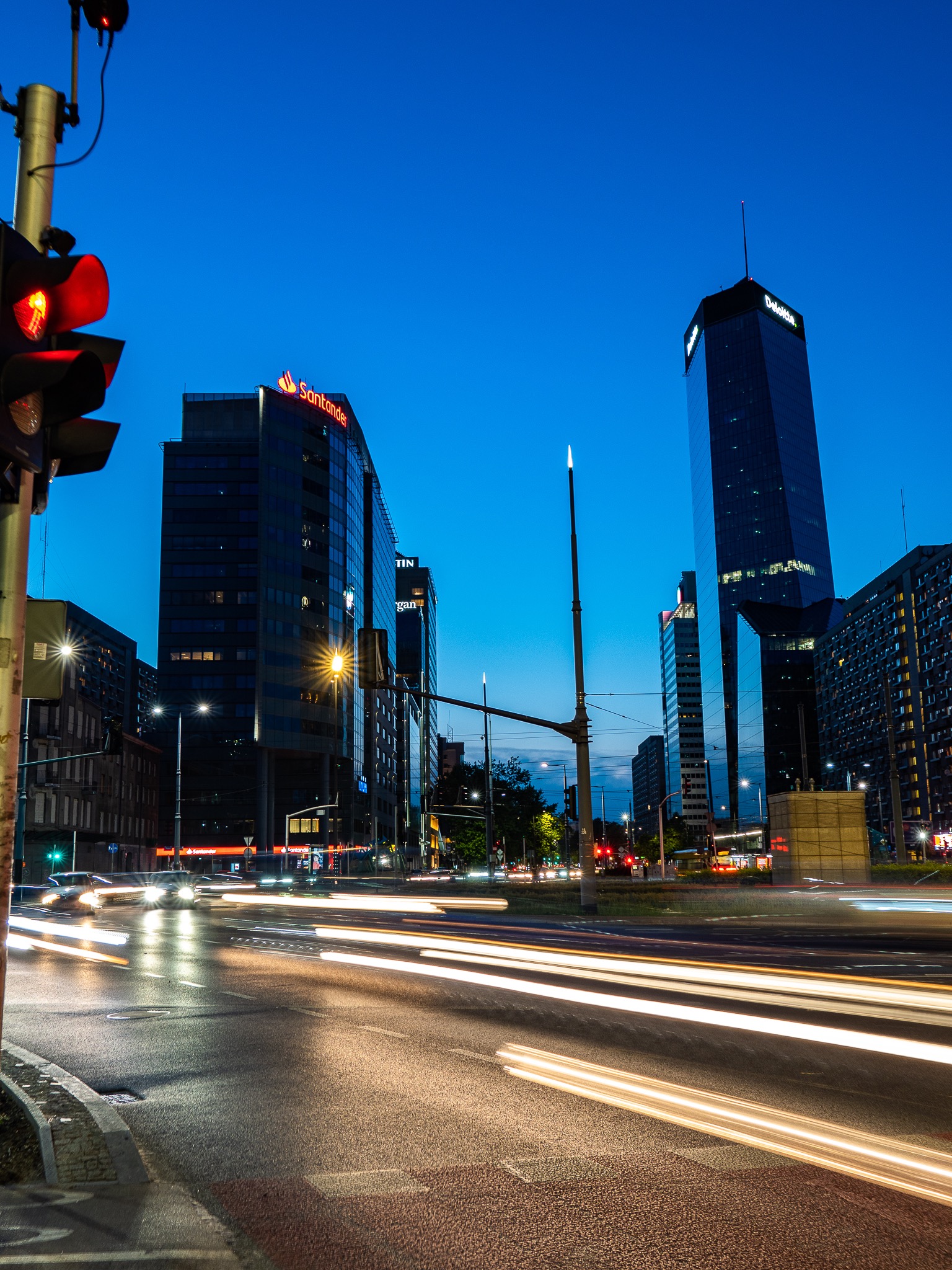 Nocny widok na miasto z wieżowcami Santander i Deloitte, światła samochodów tworzą smugi na jezdni, czerwone światło sygnalizacji.