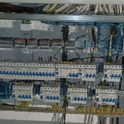 Rozdzielnica elektryczna z zainstalowanymi bezpiecznikami, przewodami i listwami zaciskowymi w szarej obudowie, widoczne okablowanie i elementy montażowe.