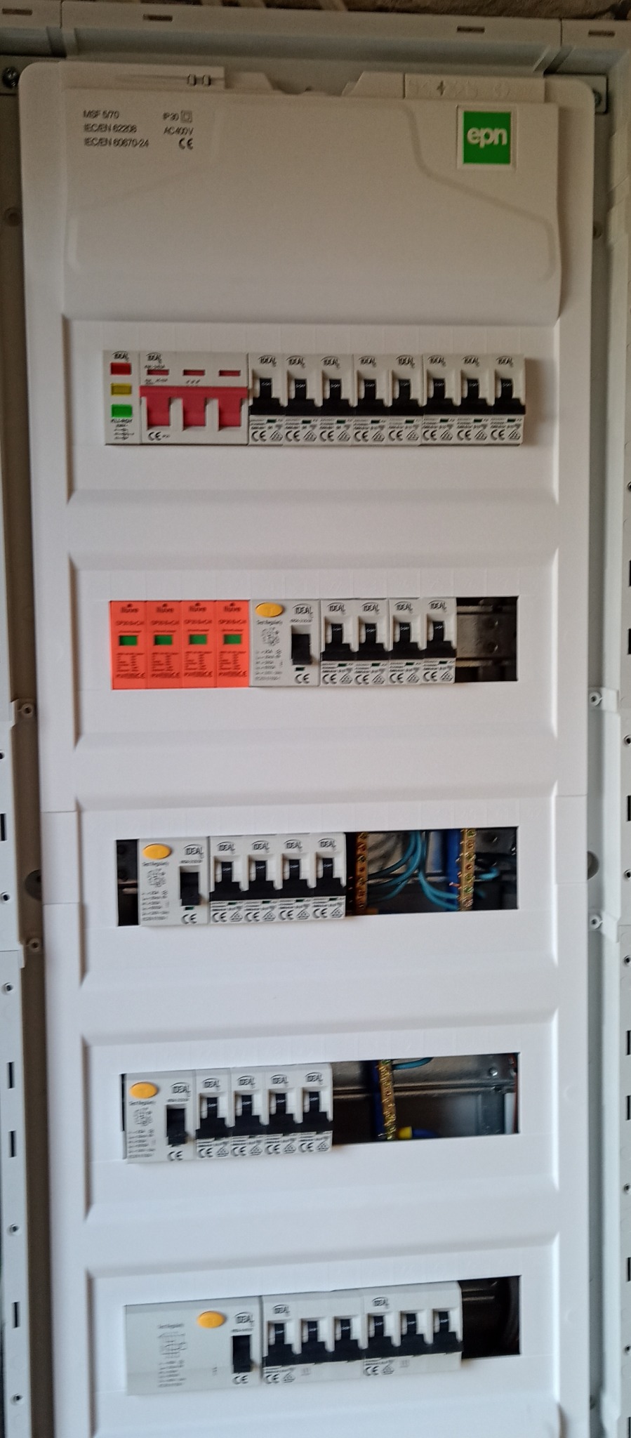 Rozdzielnica elektryczna z zainstalowanymi bezpiecznikami i wyłącznikami różnicowoprądowymi w białej obudowie. Widoczne przewody wewnątrz. Na górze logo producenta EPN.
