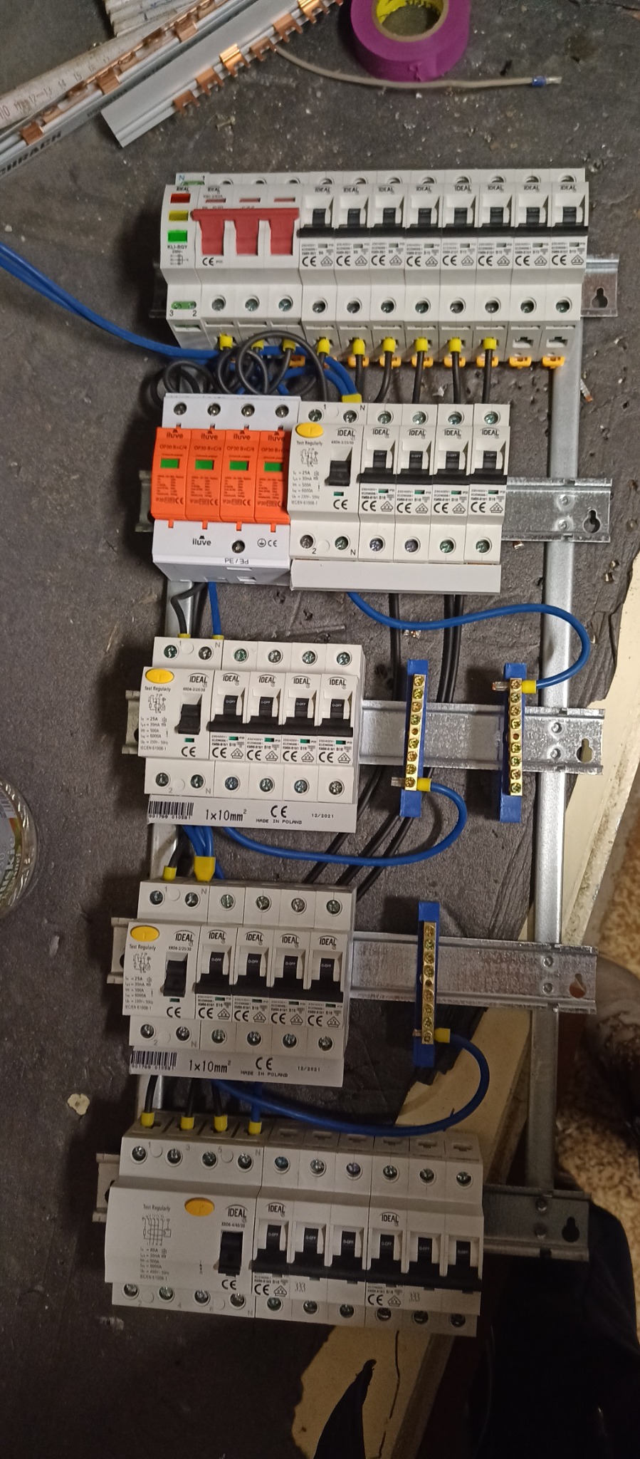 Rozdzielnica elektryczna z zabezpieczeniami różnicowoprądowymi, ogranicznikami przepięć i wyłącznikami nadprądowymi marki Ideal i Iluve zamontowana na szynie TH, widoczne niebieskie przewody...