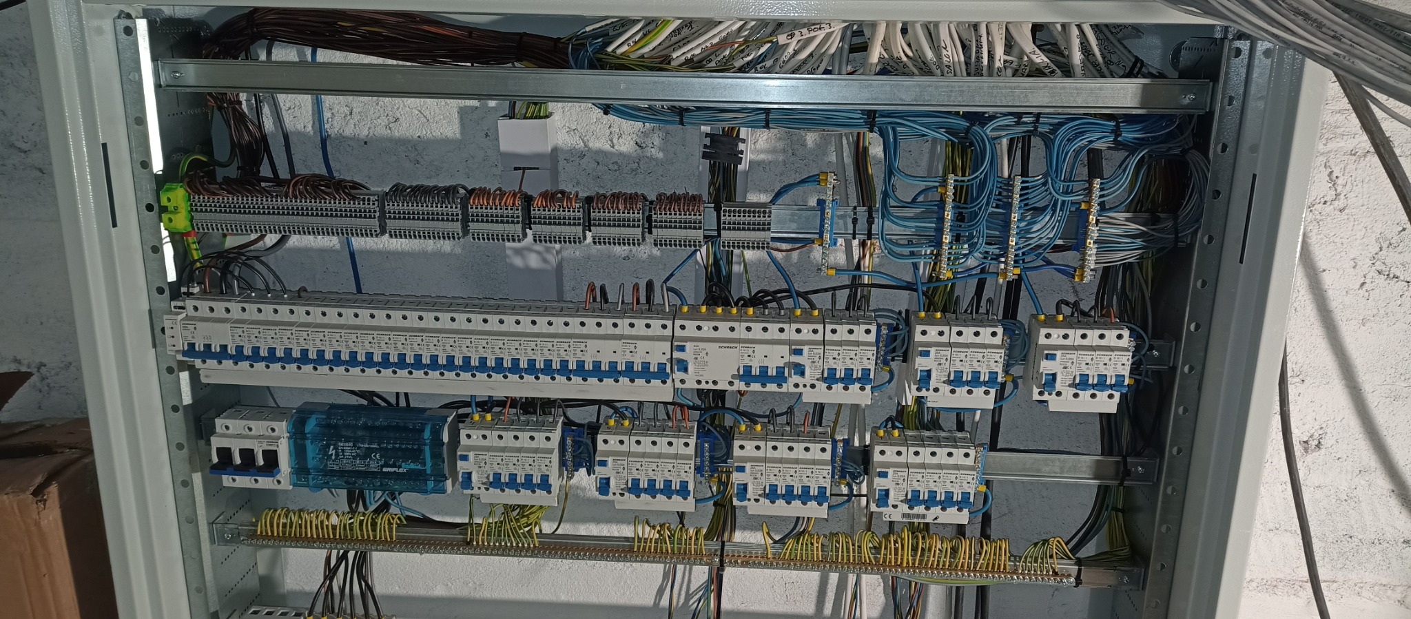 Rozdzielnica elektryczna z zainstalowanymi bezpiecznikami, przewodami i listwami zaciskowymi w szarej obudowie, widoczne okablowanie i elementy montażowe.