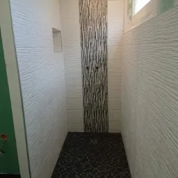 Wąska kabina prysznicowa wykończona białymi, poziomymi płytkami o fakturze oraz pionowym pasem dekoracyjnej mozaiki w odcieniach czerni i bieli, z czarną mozaiką na podłodze.