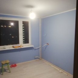 adam Szczyrba - Pokój po świeżym pomalowaniu na niebiesko, widoczne narzędzia malarskie na parapecie, odsłonięte kable w ścianie i fragmenty listew przypodłogowych na podłodze z paneli.