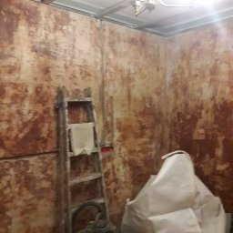 adam Szczyrba - Pokój w trakcie przygotowania do prac wykończeniowych: ściany z resztkami starej farby, drabina z szmatką, odkurzacz przemysłowy i worek z materiałem budowlanym.