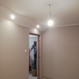 adam Szczyrba - Pomieszczenie po odświeżeniu ścian w odcieniach beżu, widoczne panele podłogowe, listwa przypodłogowa i sufit z oświetleniem punktowym, w tle mężczyzna z wiadrem i narzędziami w przejściu do innego...