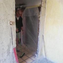 adam Szczyrba - Pracownik sprząta podłogę w pomieszczeniu w trakcie remontu, widoczny poziomica przy ścianie i folia zabezpieczająca w przejściu.