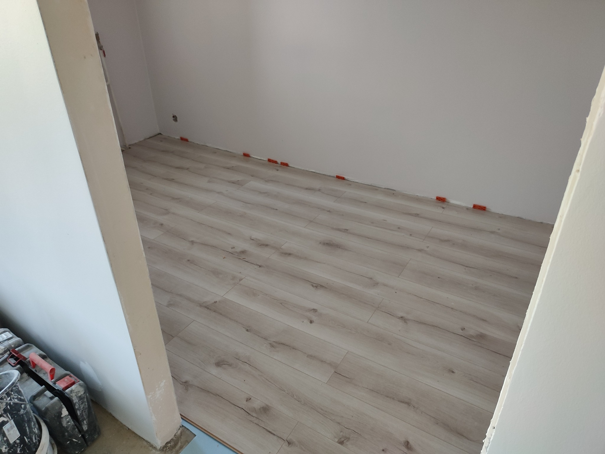 Jasne panele podłogowe ułożone w pokoju, widoczne pomarańczowe kliny dystansowe przy ścianie, w tle białe ściany i narzędzia pracy.