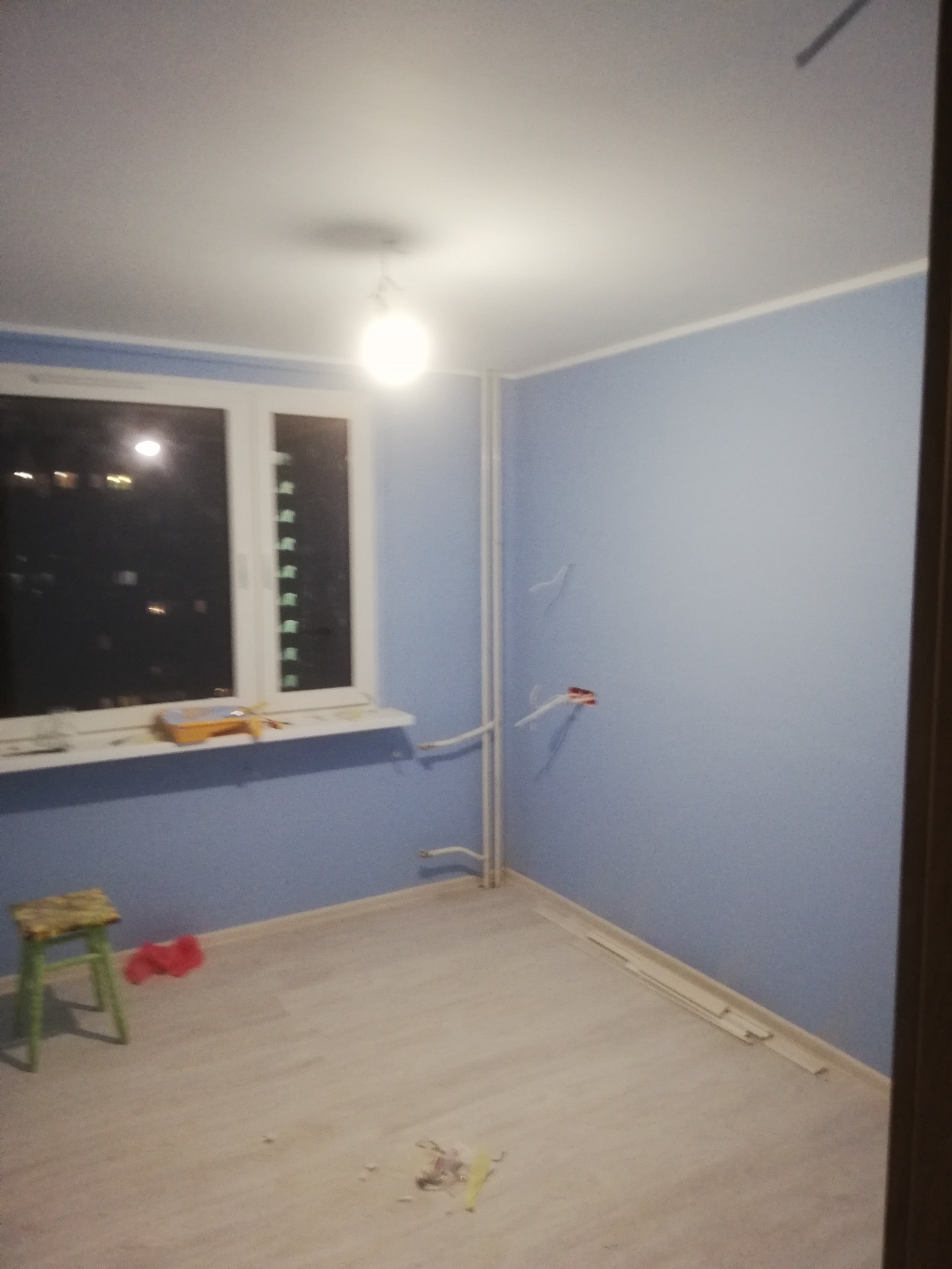 Pokój po świeżym pomalowaniu na niebiesko, widoczne narzędzia malarskie na parapecie, odsłonięte kable w ścianie i fragmenty listew przypodłogowych na podłodze z paneli.