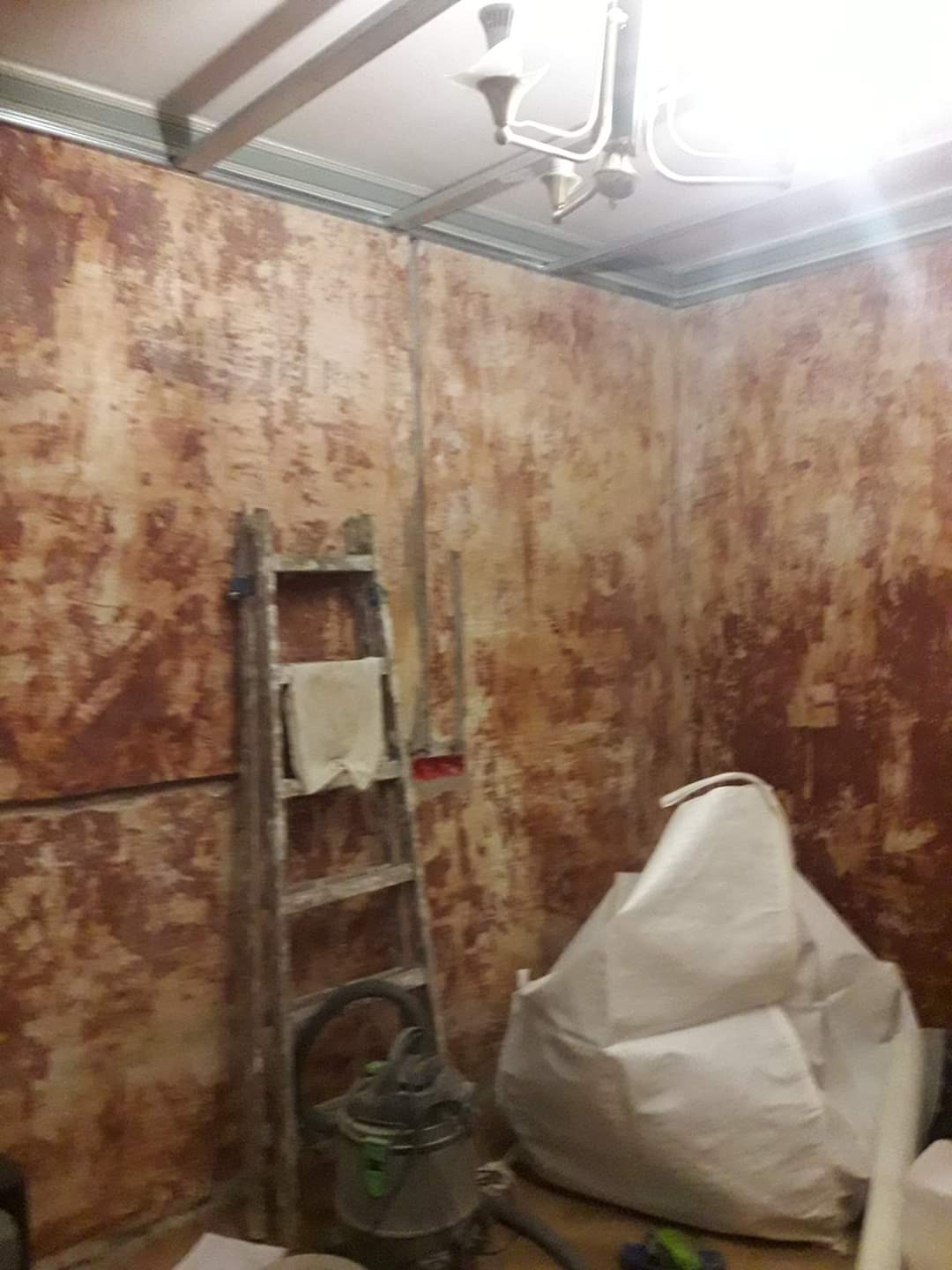 Pokój w trakcie przygotowania do prac wykończeniowych: ściany z resztkami starej farby, drabina z szmatką, odkurzacz przemysłowy i worek z materiałem budowlanym.