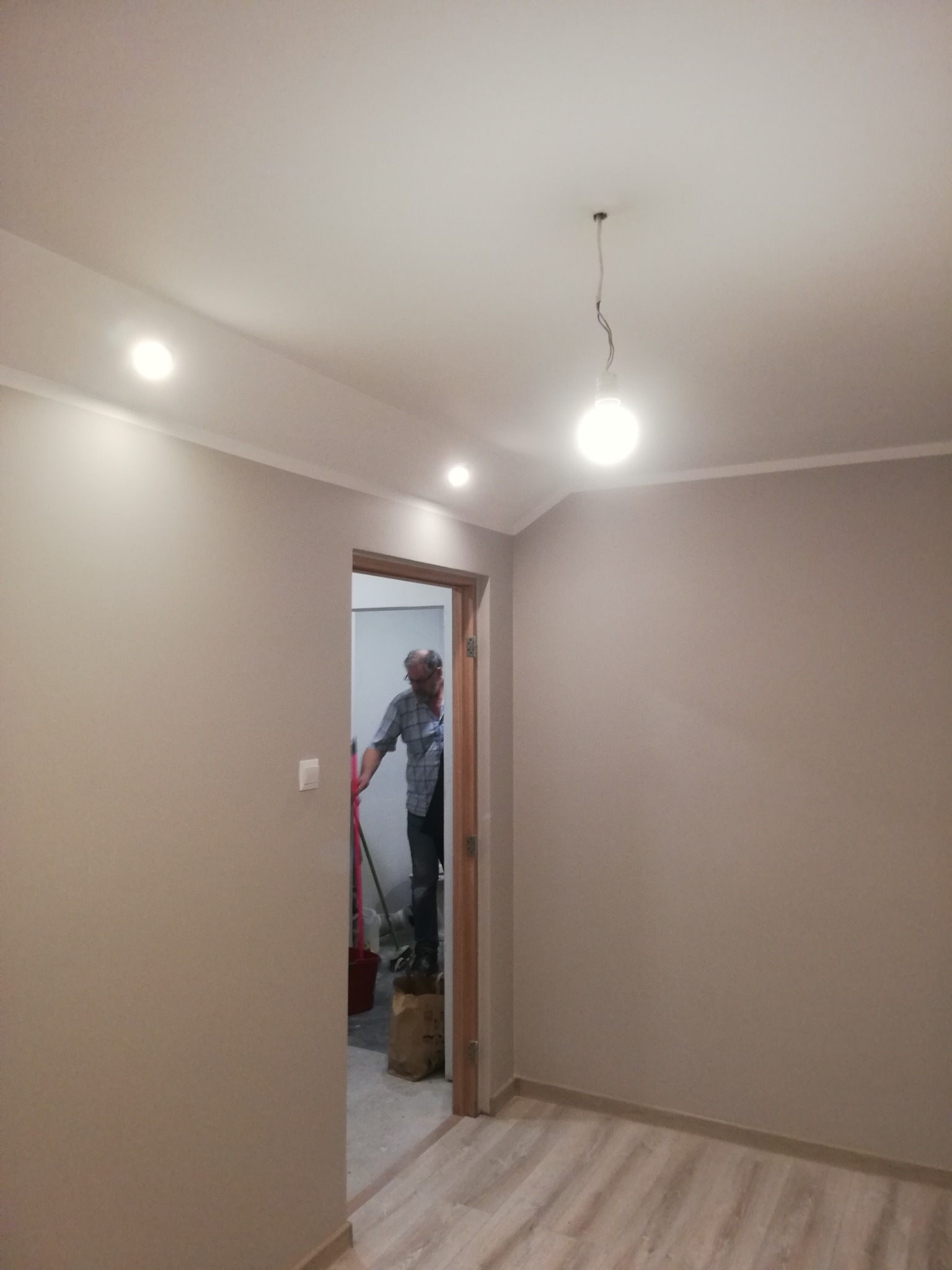 Pomieszczenie po odświeżeniu ścian w odcieniach beżu, widoczne panele podłogowe, listwa przypodłogowa i sufit z oświetleniem punktowym, w tle mężczyzna z wiadrem i narzędziami w przejściu do innego...