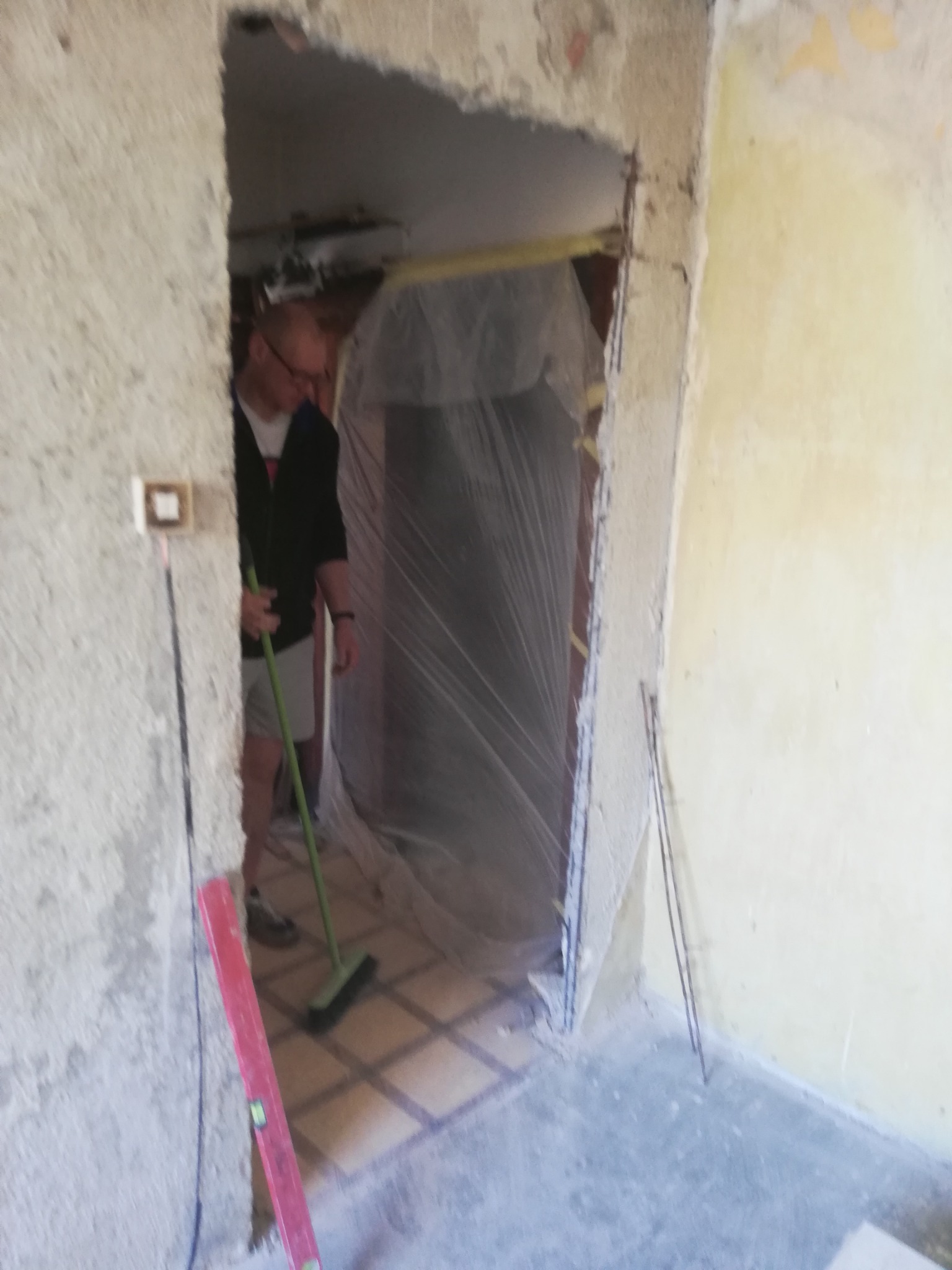 Pracownik sprząta podłogę w pomieszczeniu w trakcie remontu, widoczny poziomica przy ścianie i folia zabezpieczająca w przejściu.